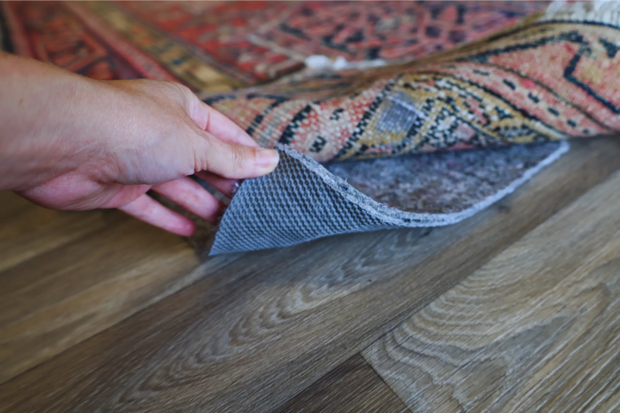 A close-up of a hand lifting the corner of a traditional oriental rug to reveal a non-slip rug pad underneath, placed on a hardwood floor for added grip and floor protection.